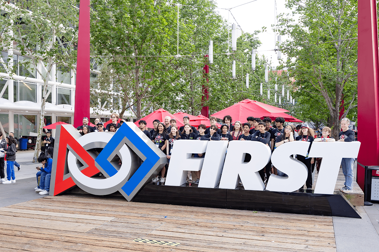 FIRST team poses for a photo behind a 3-D FIRST logo sign at FIRST Championship